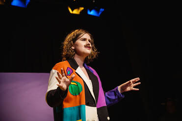 A woman wearing a multi-coloured blazer and a mustache teaches the audience. 
