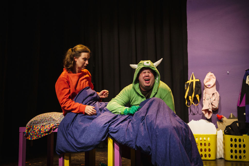 A man in a green monster suit cries over the feet of a woman in a red sweater. The woman is sitting in bed and looks at him in surprise.