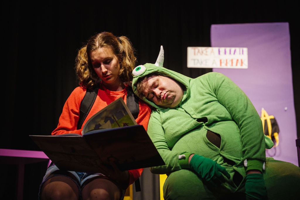 A girl in a red sweater reads a kid's book while a man in a green monster suit rests his head on her shoulder