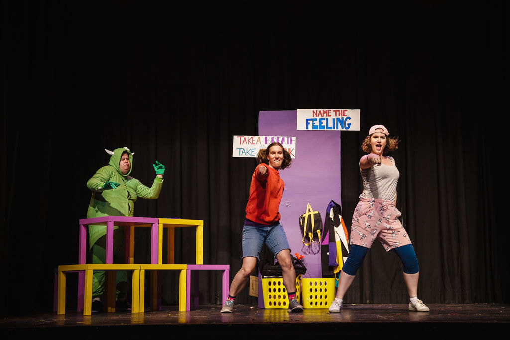 A man in a green monster suit dances behind a stack of purple and yellow tables; a girl in a red sweater and a girl in a pink hat and shorts do dance choreography together